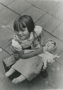 876872 Portret van Rietje Gijsen met een grote pop in de armen, zittend op een groentekistje vermoedelijk op de ...